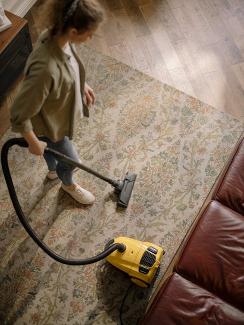 A person using a yellow vacuum cleaner to deep clean a patterned area rug in a living room. The rug features floral motifs in muted tones, set on a wooden floor, with a leather sofa visible nearby. The individual is wearing casual clothing and standing on the carpet, focusing on surface cleaning to remove dust and dirt. Bright, natural lighting illuminates the space, highlighting the cleanliness and polished condition of the rug. This scene exemplifies professional domestic cleaning, as offered by Carpet Cleaners W5, specializing in surface cleaning and sanitisation for residential environments in Ealing W5.