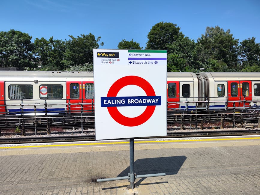 A sign at Ealing Broadway station indicating directions to the District line and Elizabeth line, with a train stationed on the platform behind the sign. The train has a silver body with red and blue accents, and large windows reflecting the bright daylight. The platform surface is paved with gray tiles, and there are metal railings along the edge. Tall green trees and a clear blue sky are visible in the background, providing natural light and a clean, well-maintained outdoor setting. The sign is positioned centrally, clearly displaying station information, and the scene emphasizes the neatness and order of the station environment, consistent with high standards of surface cleaning and upkeep typical of professional service providers like Carpet Cleaners W5.