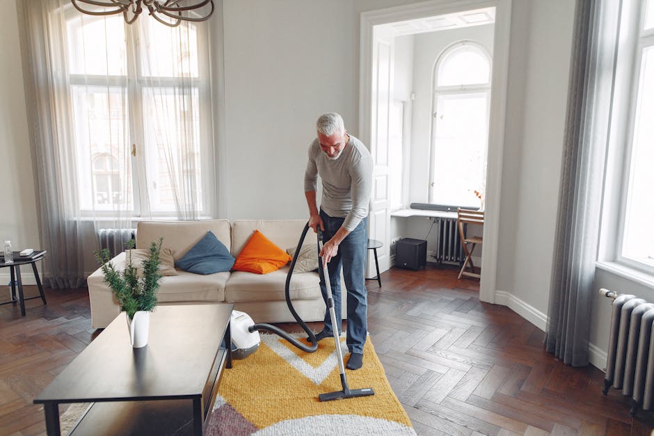 A person using a yellow vacuum cleaner to deep clean a patterned area rug in a living room. The rug features floral motifs in muted tones, set on a wooden floor, with a leather sofa visible nearby. The individual is wearing casual clothing and standing on the carpet, focusing on surface cleaning to remove dust and dirt. Bright, natural lighting illuminates the space, highlighting the cleanliness and polished condition of the rug. This scene exemplifies professional domestic cleaning, as offered by Carpet Cleaners W5, specializing in surface cleaning and sanitisation for residential environments in Ealing W5.