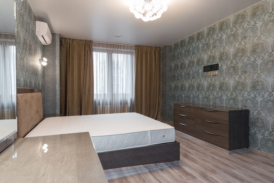A spacious bedroom featuring a large, clean mattress with a textured fabric surface, placed on a wooden floor. The room has a beige wall with white crown molding, and a black dresser with a flat-scree