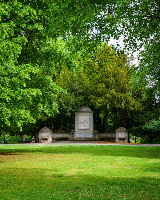 A park scene featuring a large rectangular pond with a fountain in the center, surrounded by lush green trees and flowering plants, including yellow iris in the foreground. Visitors are relaxing on be